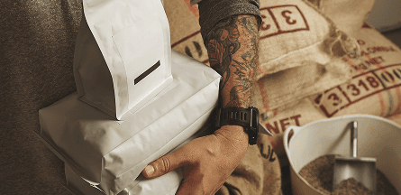 tattooed barista arm holds bags of coffee in front of burlap coffee bags
