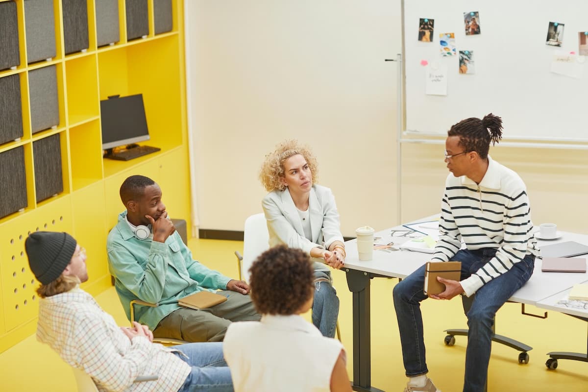 Five colleagues sit in a small group around a table, listening and reflecting during a team debrief discussion.
